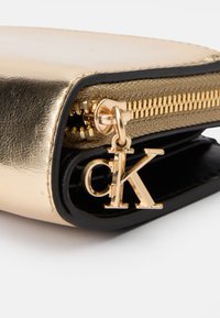 Porte-monnaie métallique doré avec des accents noirs, doté d'une fermeture éclair dorée et d'un pendentif en or en forme de 'CK' sur la tirette, présentant un design élégant et poli.
