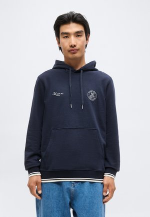 Junger Mann mit schwarzen Haaren, der einen dunkelblauen Hoodie mit weißen Logos und hellblaue Jeans vor einem einfarbigen hellen Hintergrund trägt.