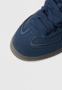 Granatowy tenisówka z miękką cholewką z zamszu, teksturowaną gumową podeszwą i usztywnionym sznurowaniem. Ma gładkie akcenty wzdłuż boków.