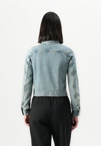 Oseba s ramenskimi dolgimi temnimi lasmi, oblečena v svetlo modro jeans jakno in črne hlače, obrnjena stran od kamere proti enobarvni beli podlagi.