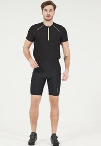 Maillot de cyclisme noir à manches courtes, avec un col zippé en néon jaune ; associé à un cuissard de compression noir et des chaussures de cyclisme.