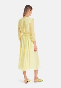 Gelbes Midi-Kleid mit durchsichtigen langen Ärmeln und tailliertem Bund. Verziert mit Blumenstickereien an den Ärmeln und einem lockeren Schnitt. Kombiniert mit grünen Ballerinas.
