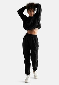 Felpa nera corta con maniche lunghe e colletto alto, abbinata a pantaloni jogger neri con vita elastica e sneakers bianche. Texture del tessuto liscia.