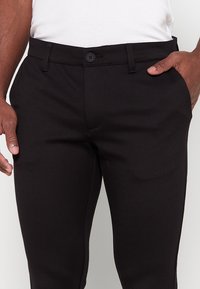 Zwarte broek van gladde stof; voorzien van een knoopsluiting en riemlussen. Tailored ontwerp met zijzakken en een slim fit silhouet.