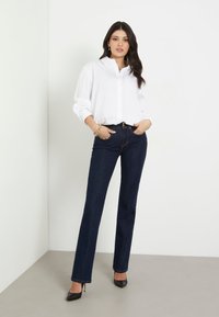 Camicia bianca con bottoni e maniche lunghe abbinata a jeans dritti di un blu scuro e scarpe con tacco nere, in piedi contro uno sfondo chiaro.