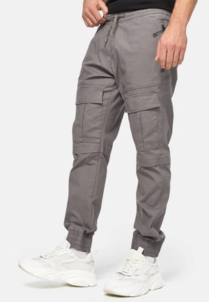 Mand iført grå cargo joggingbukser med sidelommer, elastiske manchetter og hvide chunky sneakers på hvid baggrund.