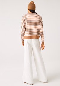 Person som bär en beige och vit randig tröja, vita vida byxor och en beige stickad mössa, stående vänd med ryggen mot en enfärgad bakgrund.