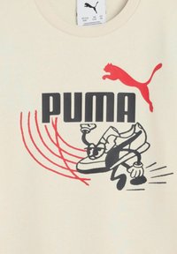 Camiseta de algodón beige con un gráfico en negro y rojo de una zapatilla, con el texto "PUMA" y un logo de puma saltando arriba.