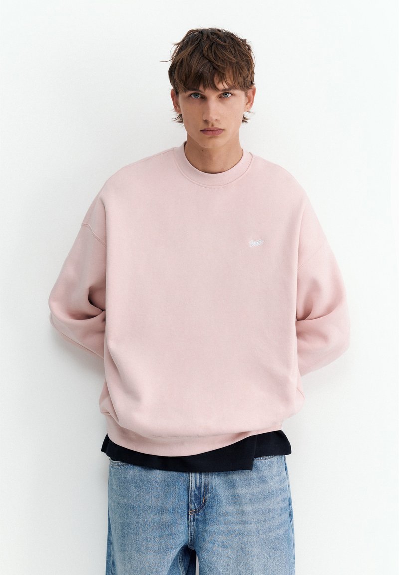Jeune homme debout, les mains dans le dos, portant un sweat-shirt rose clair sur une chemise noire et un jean bleu sur fond blanc.