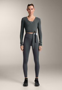 Top corto grigio a coste con maniche lunghe, caratterizzato da un design incrociato e un laccio. Abbinato a leggings grigi a vita alta e scarpe sportive nere.