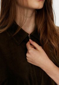 Rudas kordurojinis marškinis su apykakle ir užtrauktuku. Artimas plano vaizdas, kur matoma ranka laikanti užtrauktuką, rodanti audinio tekstūrą.