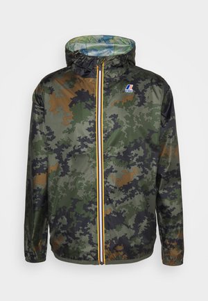 Homme portant une veste camouflage, un t-shirt noir, un pantalon blanc déchiré et des baskets blanches, debout à l'intérieur près de panneaux en verre et d'une bouche d'aération au sol.
