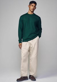 Langärmliges dunkelgrünes Hemd, cremefarbene, lockere Hose und dunkelbraune Slipper mit Metallakzenten. Weicher Stoff mit minimalistischem Design.