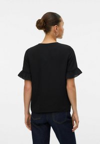 Top nero a maniche corte con polsini arricciati, realizzato in tessuto strutturato e con scollo a girocollo. Indossato con pantaloni in denim scuro.