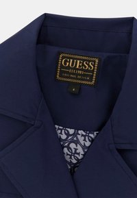 Mornarsko plava jakna s vrhunskim reverom, koja sadrži zlatnu oznaku "GUESS" i uzorčenu unutrašnjost s međusobno povezanim logotipima.