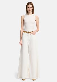 Witte geribde tanktop gecombineerd met wijd uitlopende witte broek. De outfit heeft een bruine riem en accessoires met ronde metalen accenten.