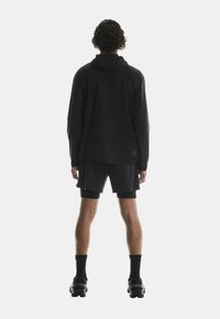 Giacca nera leggera con cappuccio, maniche lunghe e polsini elasticizzati, abbinata a pantaloni corti e scarpe sportive nere. Design semplice e funzionale.