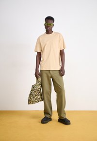 Tricou din bumbac bej cu buzunar, pantaloni verzi măslinii, pantofi slip-on negri și o geantă de voiaj cu imprimeu leopard verde. Modelul pozează pe un fundal simplu.