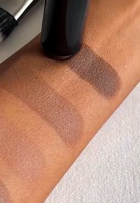 Sminkeprodukter prøvet på en solbrun arm; tre matte brune nyanser med ulik intensitet, påført med en svart applikator.