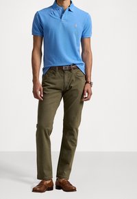 Man in een blauwe polo met korte mouwen, olivegroene broek, een bruine leren riem, bijpassende bruine leren schoenen en een polshorloge.
