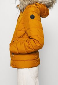 Giubbotto puffer giallo senape con cappuccio staccabile foderato in pelliccia, tasche con zip e una toppa rotonda nera sul manico sinistro.