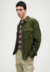 Ελαιόχρωμο κοτλέ σακάκι με γιακά και δύο τσέπες στο στήθος, φορεμένο πάνω από πουκάμισο με καρό σχέδιο και συνδυασμένο με χακί cargo παντελόνι.