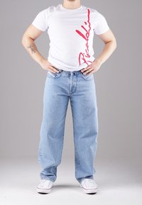 Valkoinen t-paita, jossa punainen painatus sivussa, yhdistetty vaaleansinisiin leveälahkeisiin farkkuihin ja valkoisiin tennareihin. Yksinkertainen malli ja rento istuvuus.