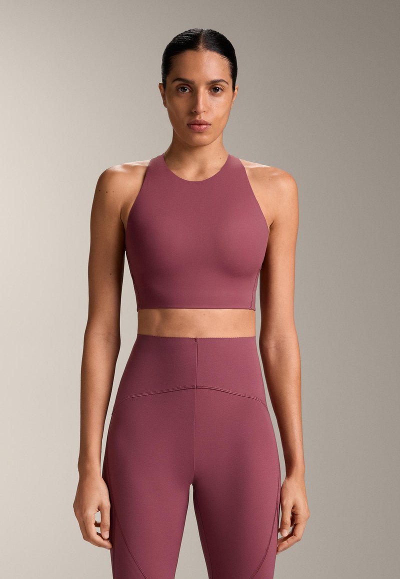 Donna in piedi di fronte che indossa un reggiseno sportivo a collo alto e pantaloni aderenti a vita alta abbinati color malva, su sfondo beige.