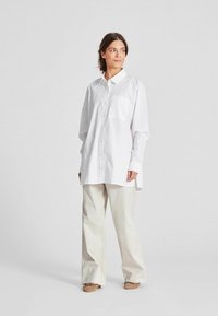 Camicia bianca oversize con taschino sul petto, maniche lunghe e un taglio morbido, abbinata a pantaloni larghi di un beige chiaro e scarpe basse.