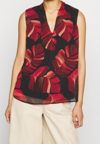 Ärmlös svart blus med röd och brun tropisk lövmönster, bärs tillsammans med lösa beige byxor.