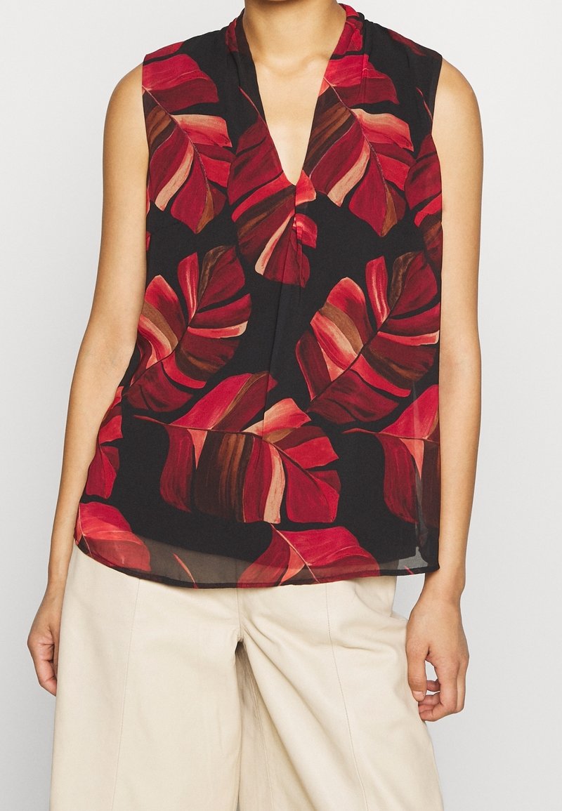 Ärmlös svart blus med röd och brun tropisk lövmönster, bärs tillsammans med lösa beige byxor.
