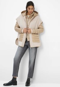 Beige gewatteerd vest met een capuchon, met een gladde buitenstof, een gestikte textuur en twee voorkruiszakken. Gekoombineerd met grijze spijkerbroek en zwarte instappers.