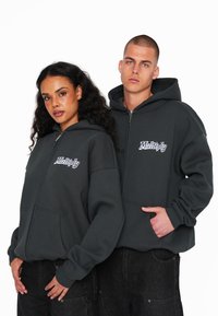 Felpe con zip in modalità charcoal, dotate di tasche frontali e testo ricamato "Multiply". Vestibilità ampia e tessuto morbido. Modello in posa ravvicinata.