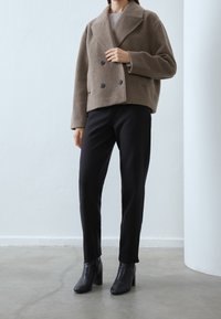 Brun dubbelknäppt kavaj i ullblandning med stora knappar, i kombination med svarta åtsittande byxor och ankelstövlar, mot en ljus bakgrund.