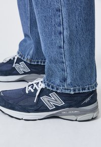 Pieds portant des baskets New Balance 990 bleu marine avec des lacets blancs et un jean en denim bleu clair.
