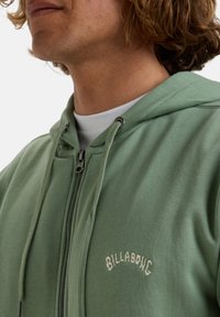Haut à zip vert en matériau doux, doté d'une capuche avec cordon de serrage et d'un logo brodé "BILLABONG" en crème sur la poitrine.