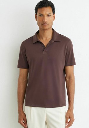 Mand står mod en hvid væg iført en brun kortærmet polo-shirt og lyse bukser, vendt fremad.