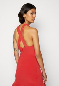 Vestido sem mangas vermelho com alças cruzadas e silhueta ajustada. Apresenta uma bainha rodada e tecido texturizado. Fecho com zíper nas costas.