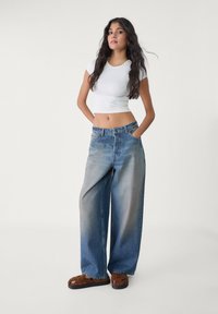 Witte cropped t-shirt, wijde vervaagde blauwe jeans, bruine instap sandalen. Model poseert met handen in de zakken tegen een neutrale achtergrond.