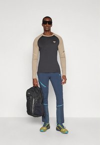Schwarz-braunes Langarmshirt, blaue Hose mit hellblauen Akzenten, schwarzer Rucksack und Wanderschuhe mit gelben Sohlen. Sonnenbrille am Modell.