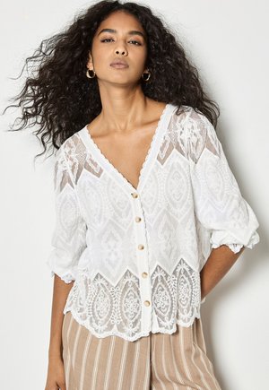 Donna con lunghi capelli ricci che indossa orecchini a cerchio dorati, una camicetta bianca di pizzo con bottoni e bordi smerlati, e pantaloni beige a righe.