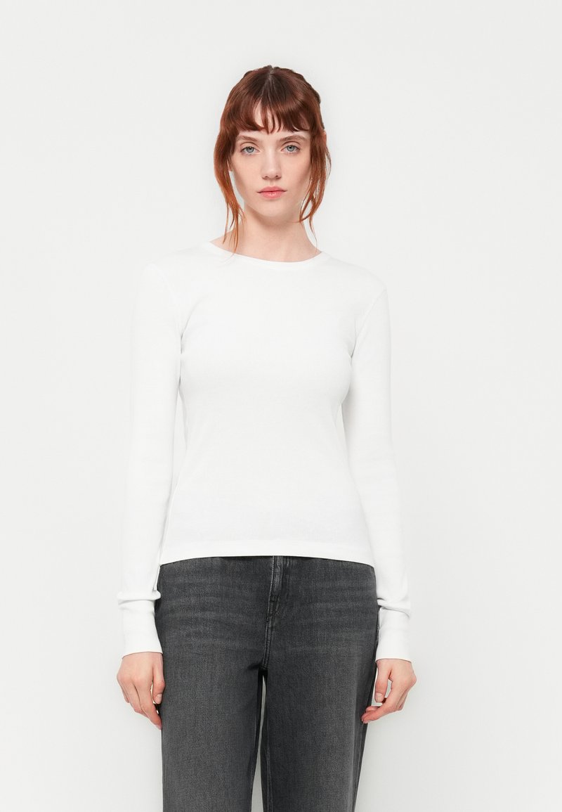 White long-sleeve shirt with a crew neckline and ribbed texture, paired with dark gray pants. Simple and minimalistic design.
