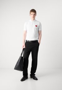 Hvid polo shirt med et rødt logo-patch, parret med sorte dressbukser. Modellen holder en sort læder tote bag og har sorte sko på.