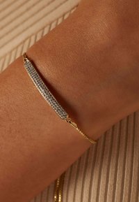 Bracelet en chaîne dorée avec une barre rectangulaire ornée de cristaux transparents, sur un fond texturé de teint de peau.