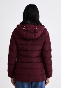 Burgundy stoppad jacka med huva, med horisontell quiltad design och tryckknappsdetaljer vid halsringning och ärmslut.