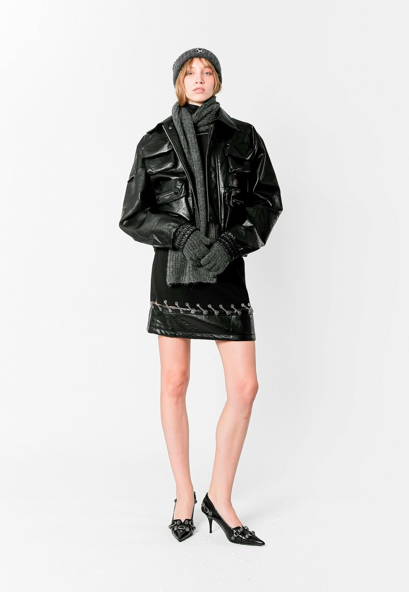 Chaqueta de cuero negra con grandes bolsillos, vestido negro con un dobladillo decorativo, bufanda de lana gris, guantes y zapatos de tacón con detalles decorativos.