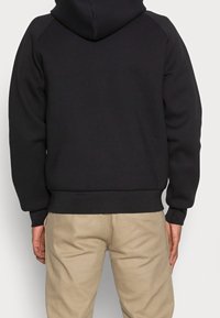 Persona che indossa una felpa nera con cappuccio e pantaloni beige, vista da dietro su uno sfondo chiaro e semplice.