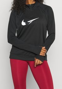 Person in schwarzem Langarm-Nike-Oberteil mit weißem Logo und Reißverschluss, trägt rote Leggings und passt die Ärmel auf Taillenhöhe an.