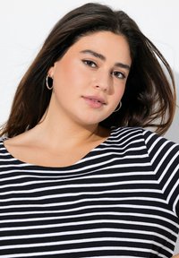 Musta-valkoinen raidallinen paita pyöreällä kaula-aukolla, lyhythihaisena ja pehmeästä materiaalista. Mallilla on pitkät hiukset ja hänellä on hopeiset renkaakorvakorut.