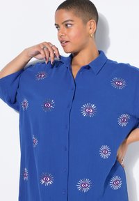 Chemise bleue à manches longues en tissu texturé, ornée de motifs d'yeux brodés avec des accents violets et des designs d'éclats métalliques.